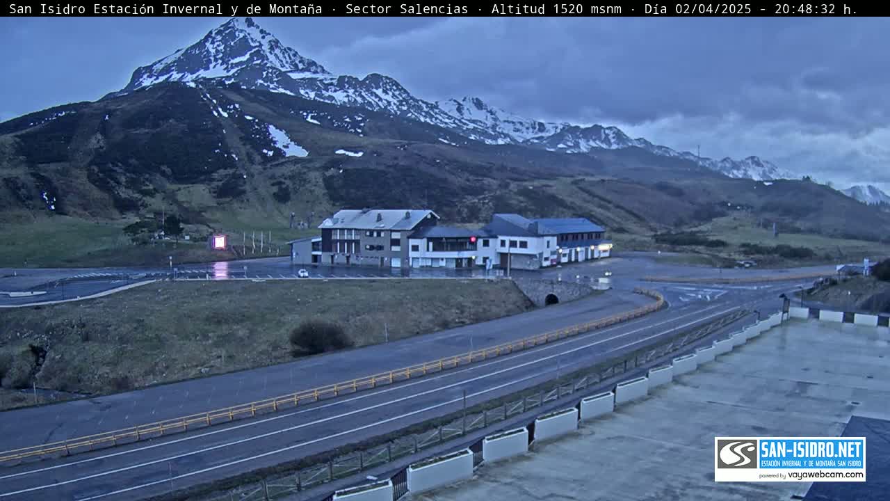A mountain range with snow-capped peaks is visible under a partly cloudy, dusky sky, overlooking a parking lot and roadway near a building complex.