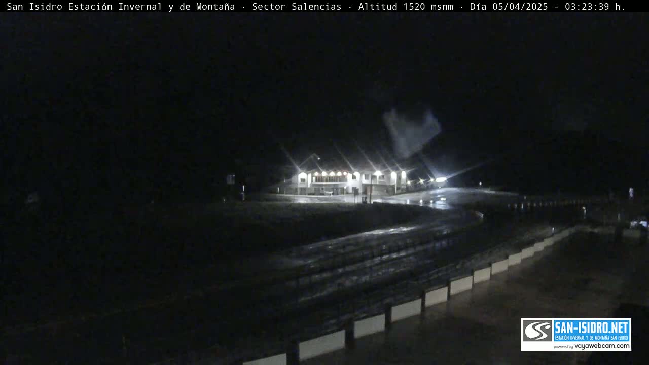 A nighttime, wet scene shows a building with lights on and a roadway with guardrails.