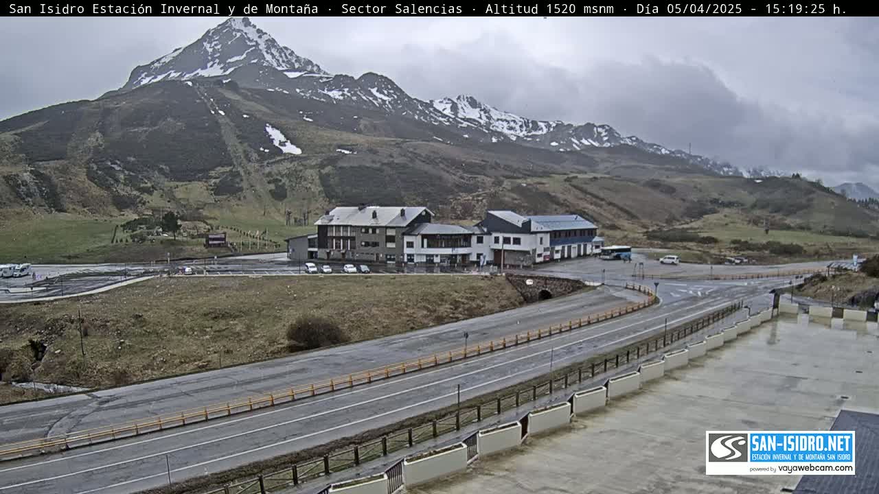 A mountain range with patches of snow is visible in the background under an overcast sky, overlooking a parking lot and road leading to a cluster of buildings near the base of the mountain.