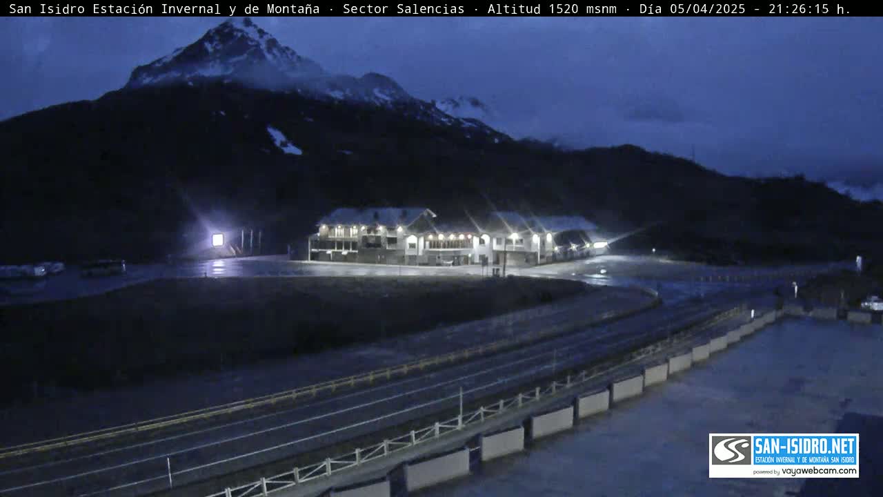 A mountain lodge is illuminated at night, nestled below a dark mountain range under a twilight sky.