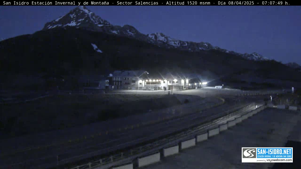 A mountainside building complex is illuminated at night under a dark, clear sky, with snow-capped mountains in the background.