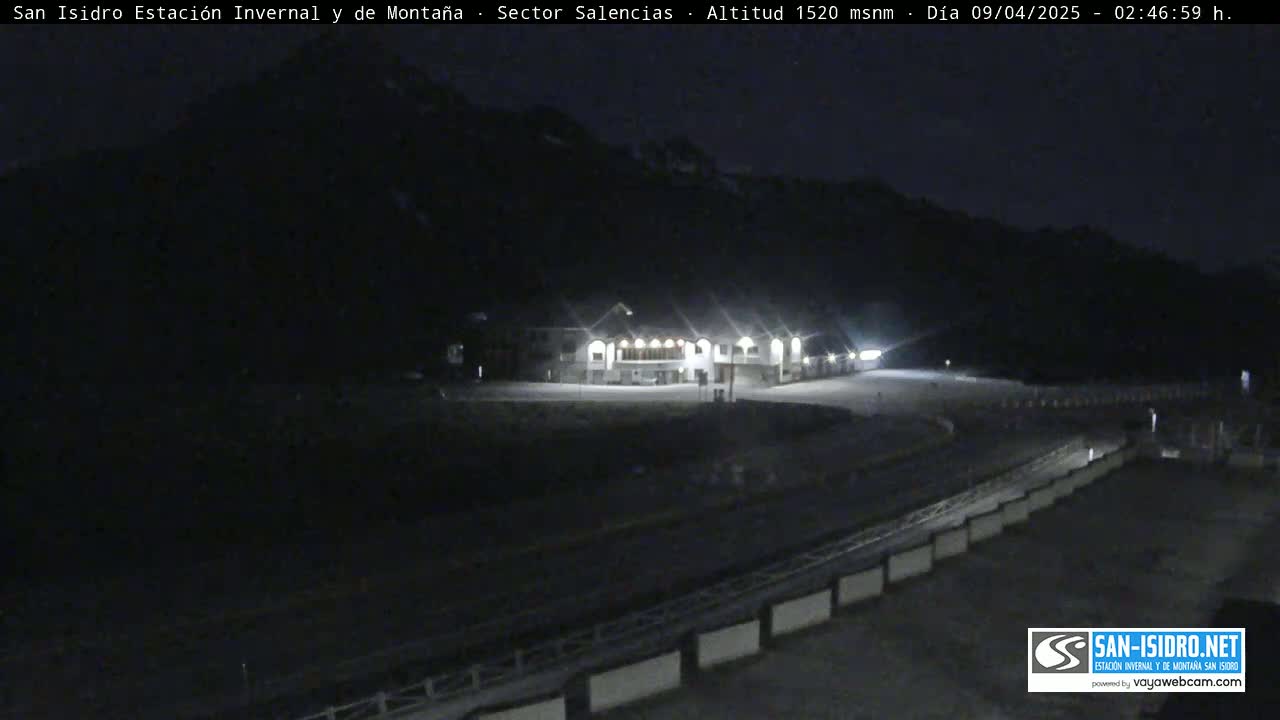 A brightly lit building sits in a dark, snow-covered area at night, with mountains in the background.