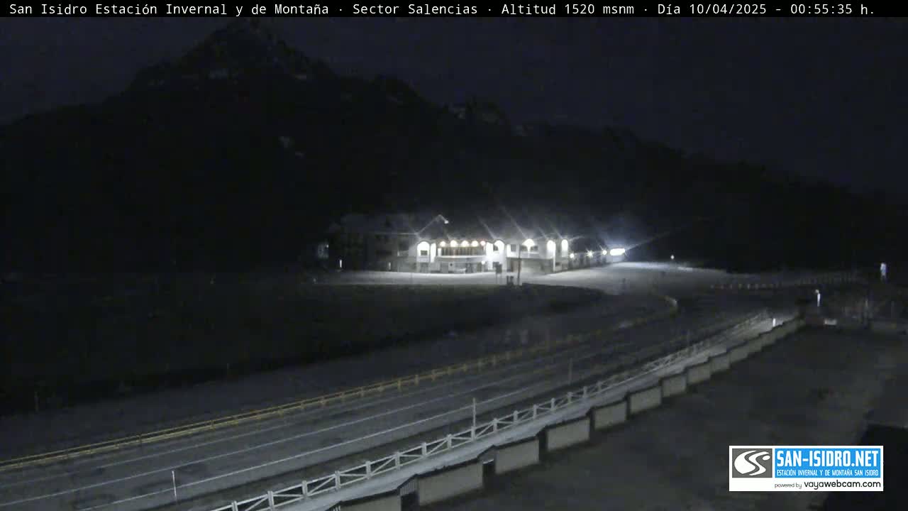 A nighttime, snow-covered mountain scene shows a brightly lit building near a roadway with barriers.
