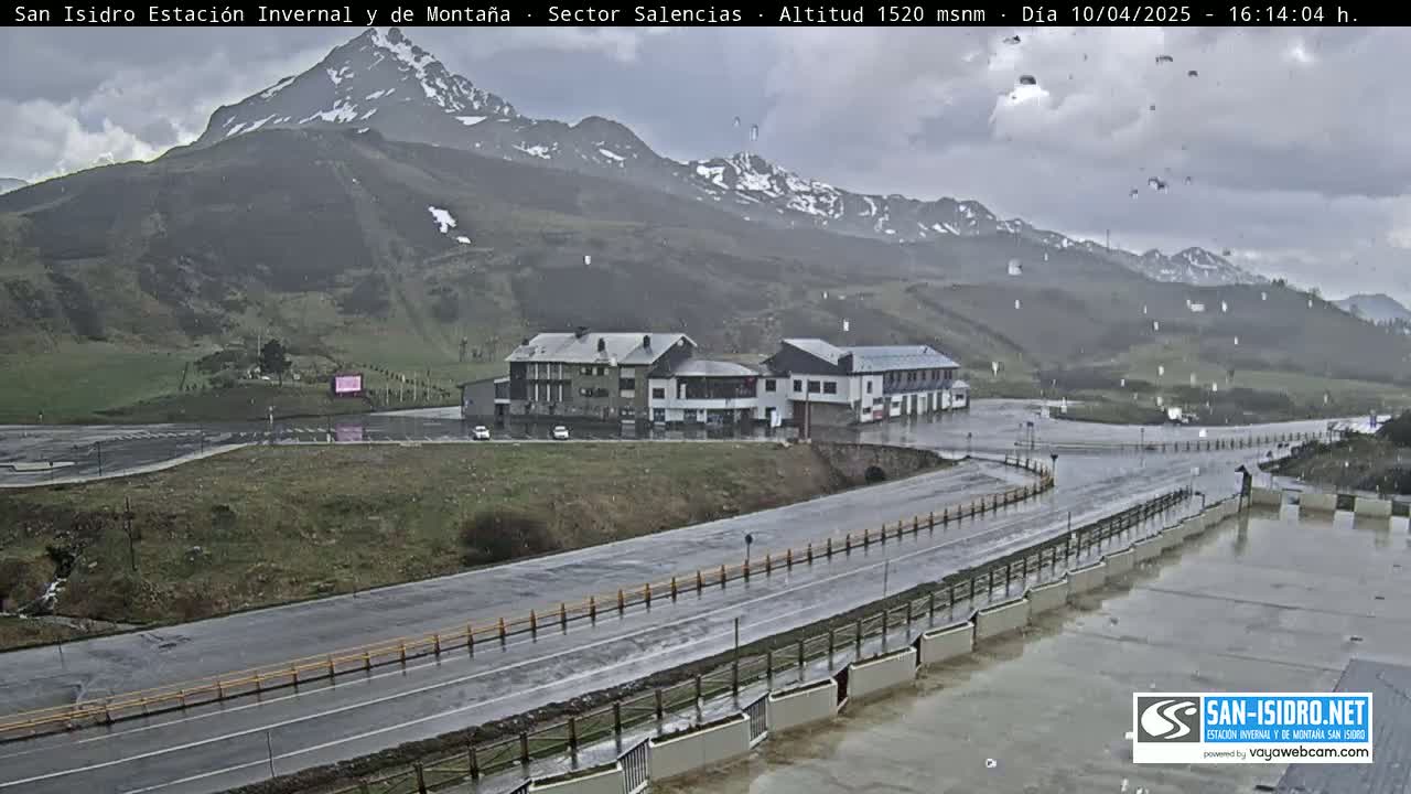 A wet, overcast mountain road curves past a building complex near a snow-dusted mountain range.