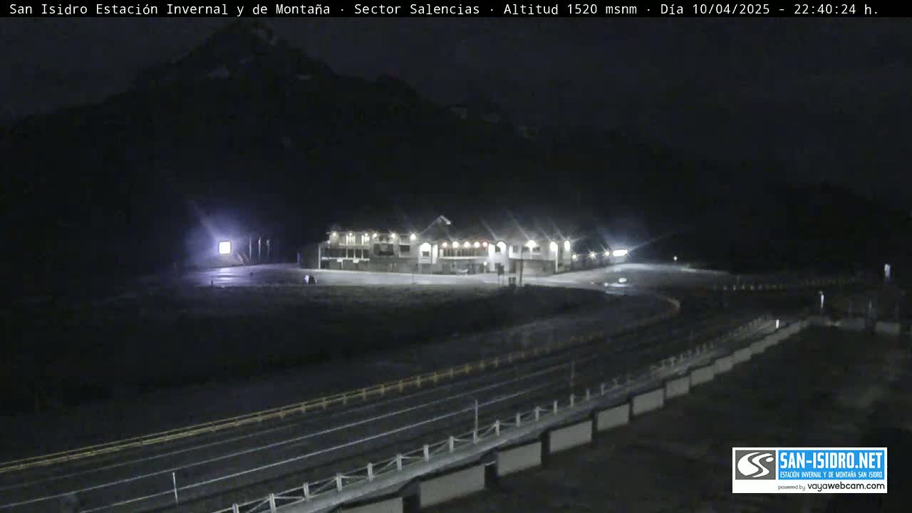 A brightly lit building complex sits near a curving road at night, with a mountain looming in the background under a dark sky.