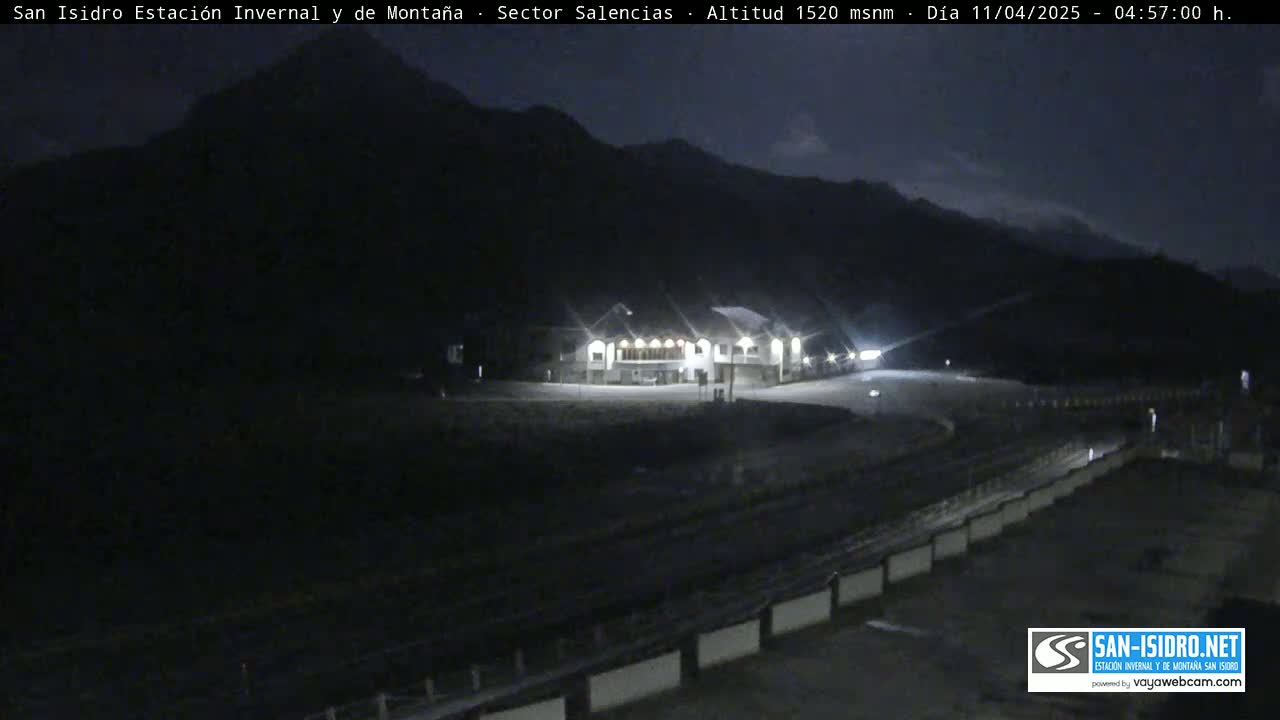 A dimly lit mountainside scene at night shows a building with lights on, a roadway curving around it, and dark mountains in the background under a dark, mostly clear sky.