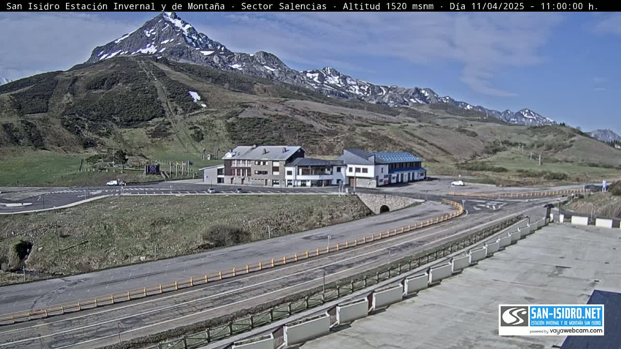 A mountainside scene on a sunny day shows a parking lot and several buildings near a roadway, with snow-capped mountains in the background.
