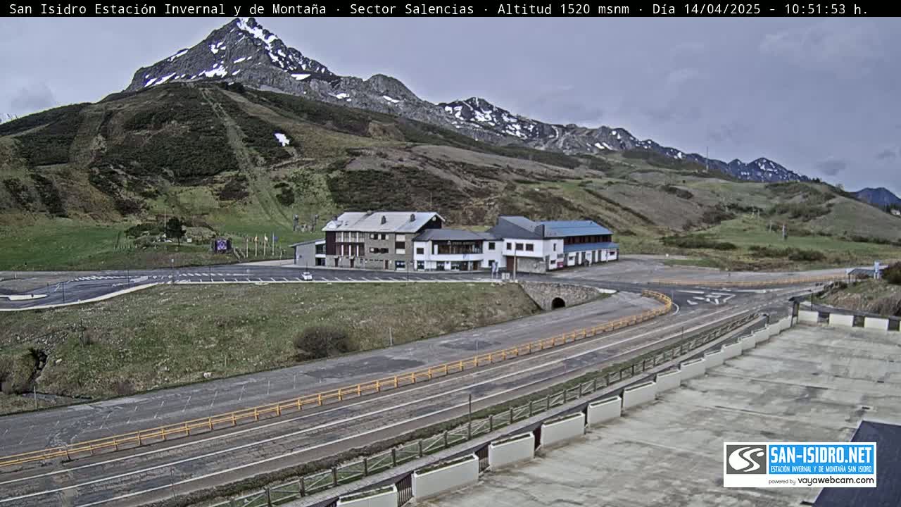 A mountain resort with a parking lot and roadway sits below snow-capped mountains under an overcast sky.
