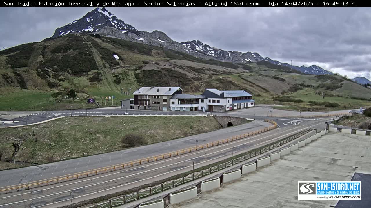 A mountainside parking lot with a few buildings is situated below a snow-capped mountain range under an overcast sky.