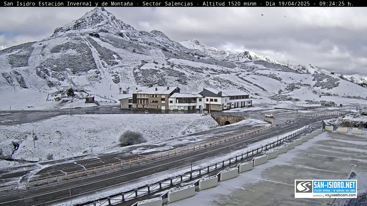 A snow-covered mountainside with a road and buildings, under an overcast sky.