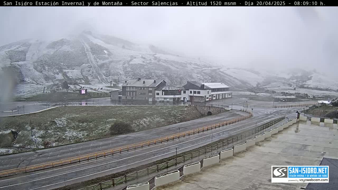 A snow-covered mountain slopes down to a road and buildings, with light snow falling in overcast conditions.