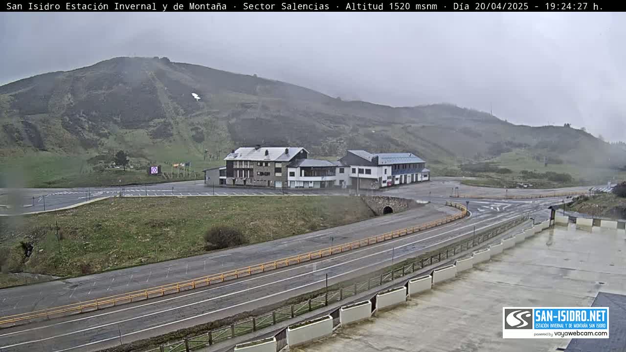 A mountainside parking lot and roadway with two buildings sit below a hill, under an overcast sky.