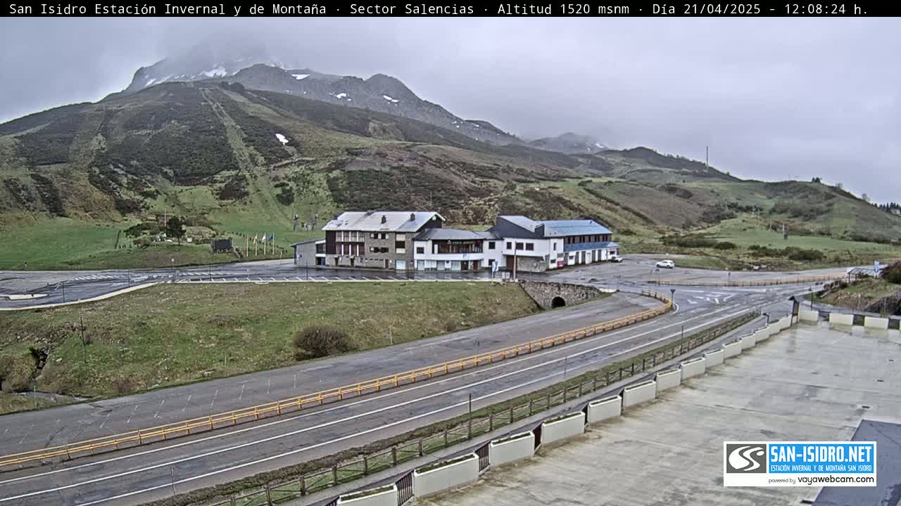 A mountainside parking lot with a few buildings sits beside a paved road on an overcast day.
