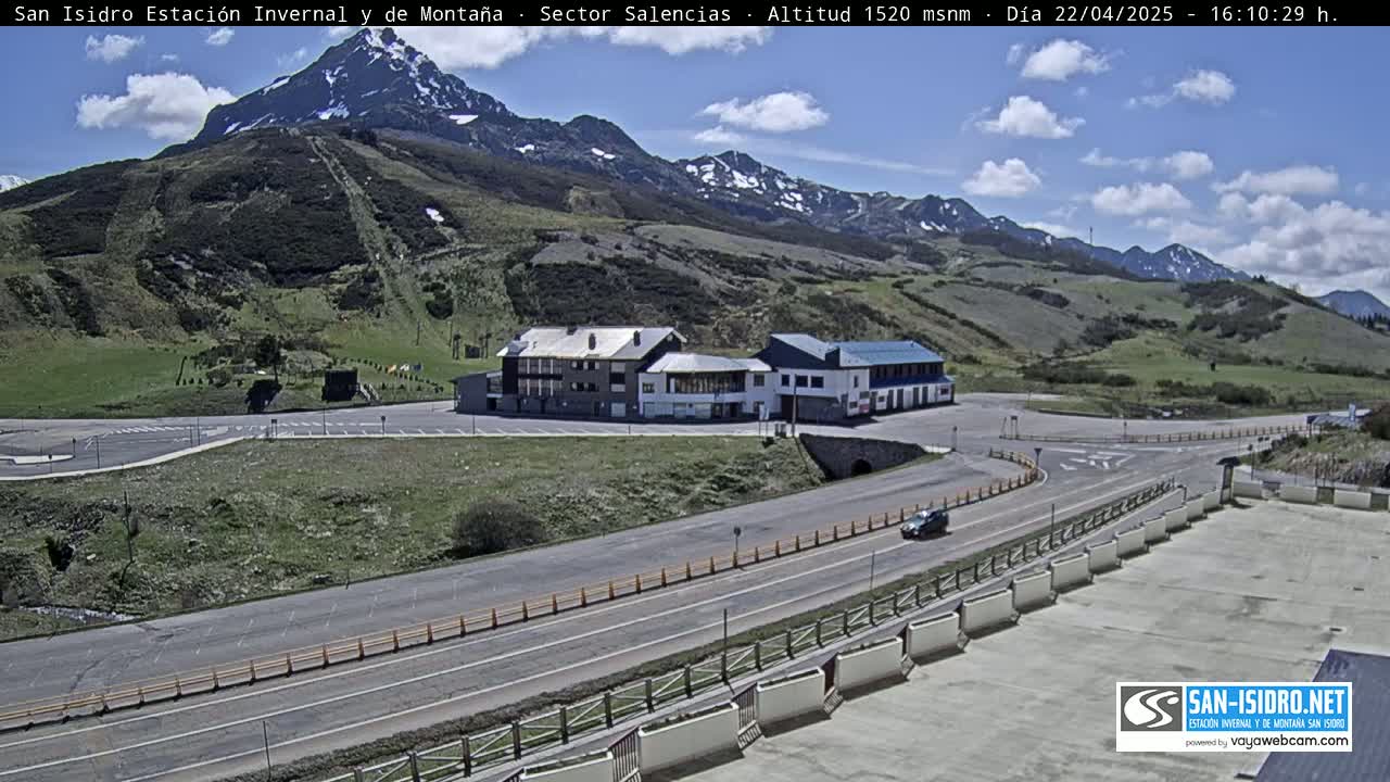 A mountain road curves past a building complex and parking lot, with a partly cloudy sky above and snow-capped mountains in the background.