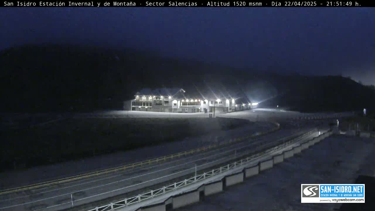 A snow-covered, nighttime scene shows a large building illuminated against a dark mountainside.