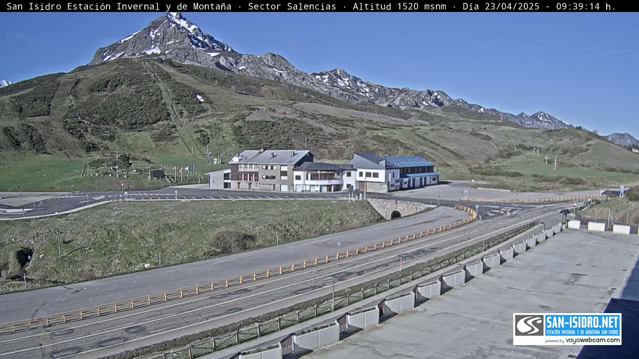 A sunny mountain scene shows a paved road curving around several buildings near a parking lot, all set against a backdrop of a snow-capped mountain.