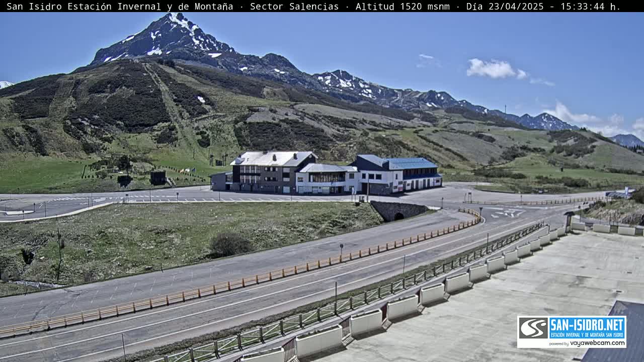 A mountain resort with a large parking lot and several buildings sits below a snow-capped mountain range under a mostly clear blue sky.