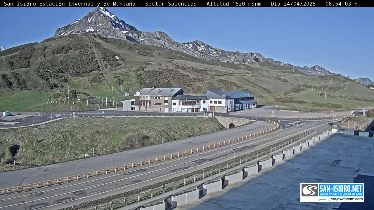 A mountainside parking lot with several buildings and a roadway leading to a mountain range under a clear blue sky.
