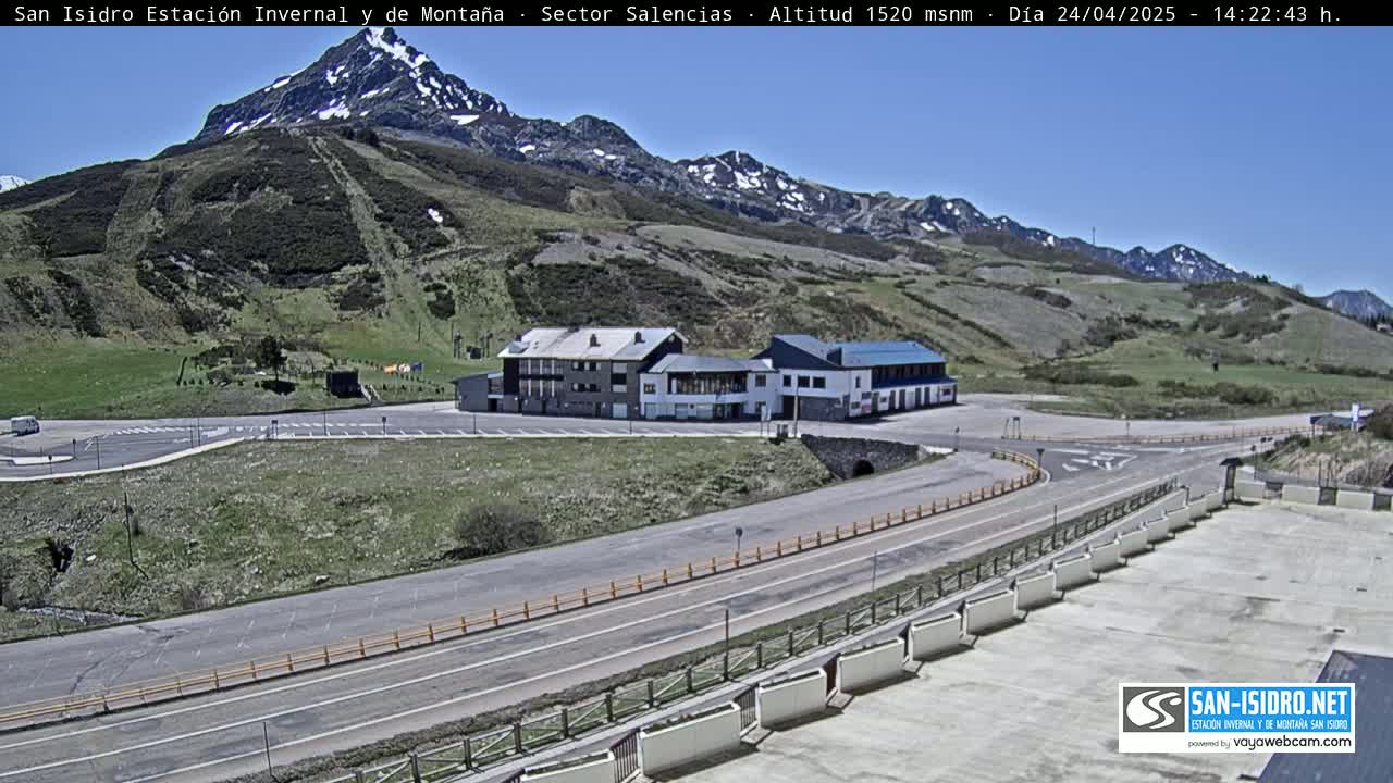A mountainside parking lot with a building complex and a road curving around a hillside, under a clear blue sky.