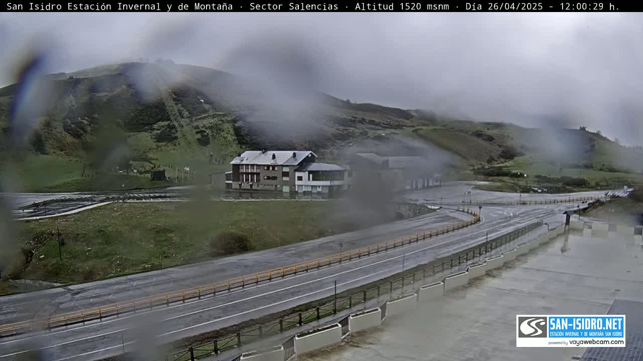 A wet, overcast view of a mountain road curving past a building complex, with a hill in the background.