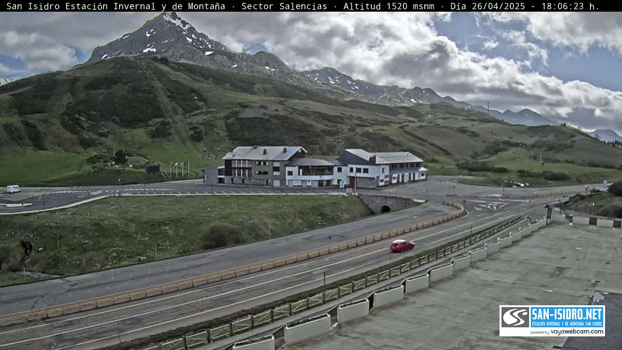 A mountainside with a building complex and parking lot is visible under a partly cloudy sky, with a road curving through the scene.
