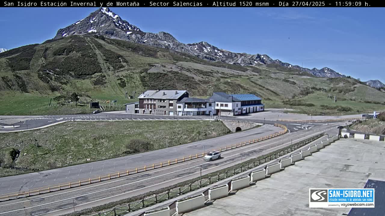 A mountain road winds through a parking lot and past a building complex, with a clear blue sky overhead and snow-capped mountains in the background.