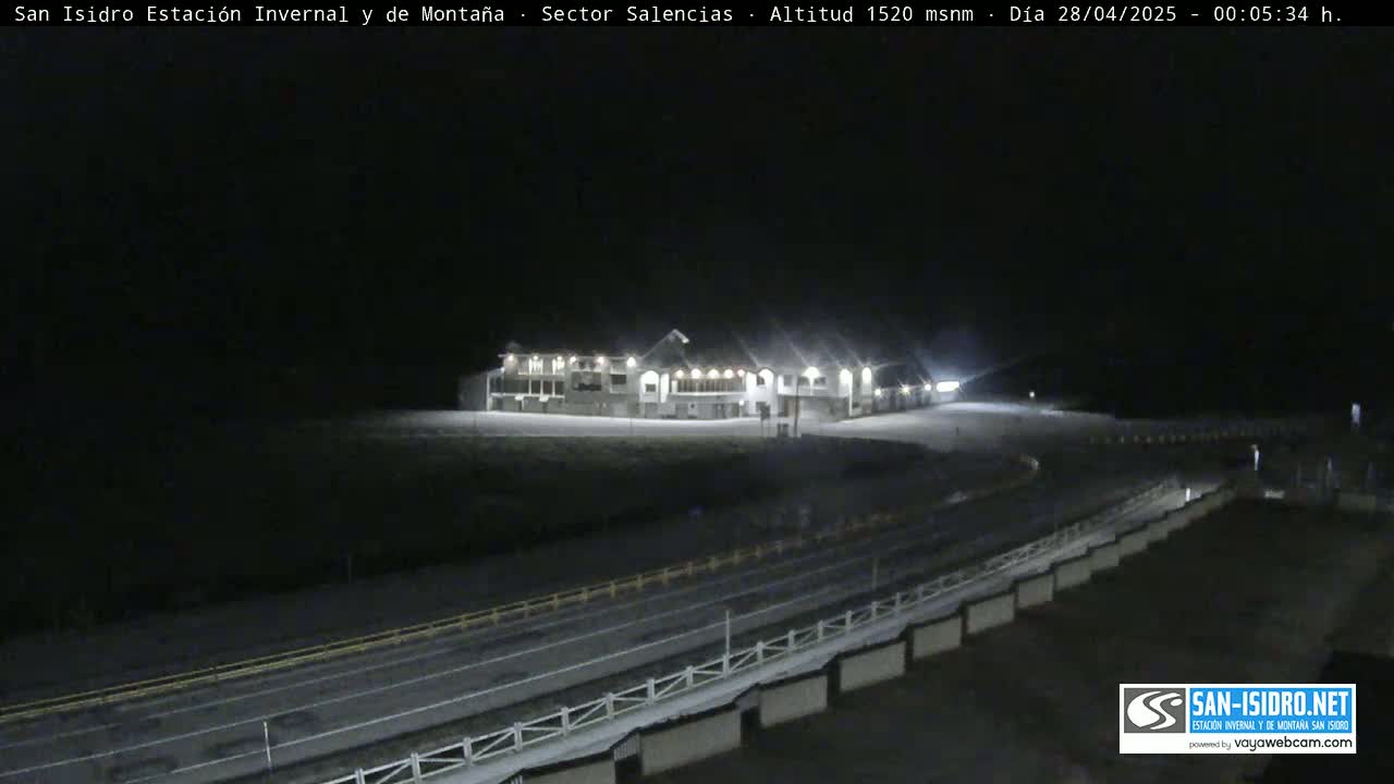 A brightly lit building sits atop a snow-covered hill at night, overlooking a road with a low barrier.