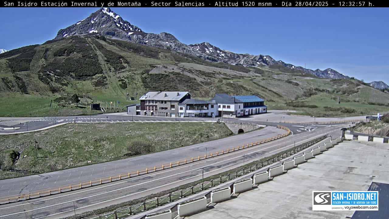A mountainside parking lot with a building complex and a road curves around a snow-capped mountain under a clear blue sky.