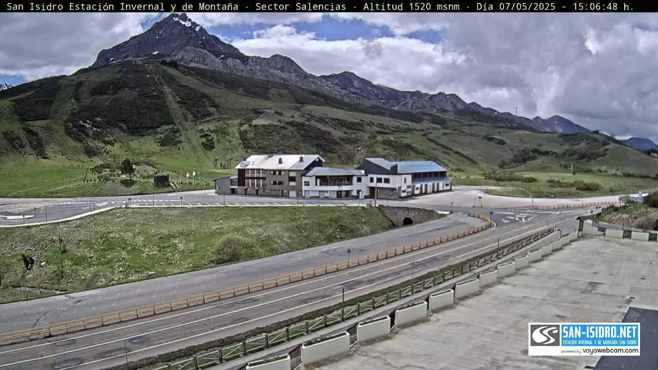 A mountainside scene with a partly cloudy sky shows a road curving past a cluster of buildings near a mountain with visible ski slopes.