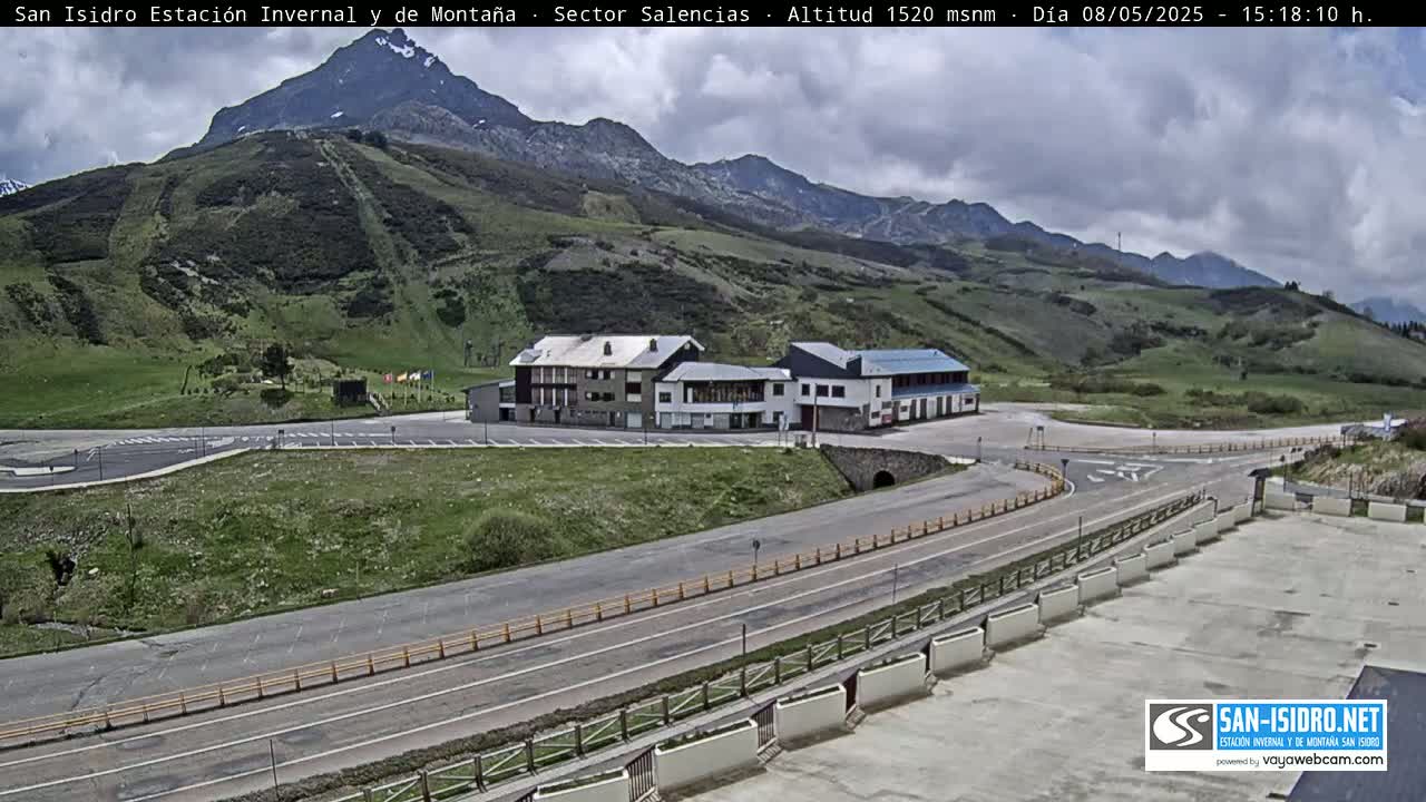 A mountainside scene under partly cloudy skies shows a roadway curving around a parking lot and several buildings near a ski slope.
