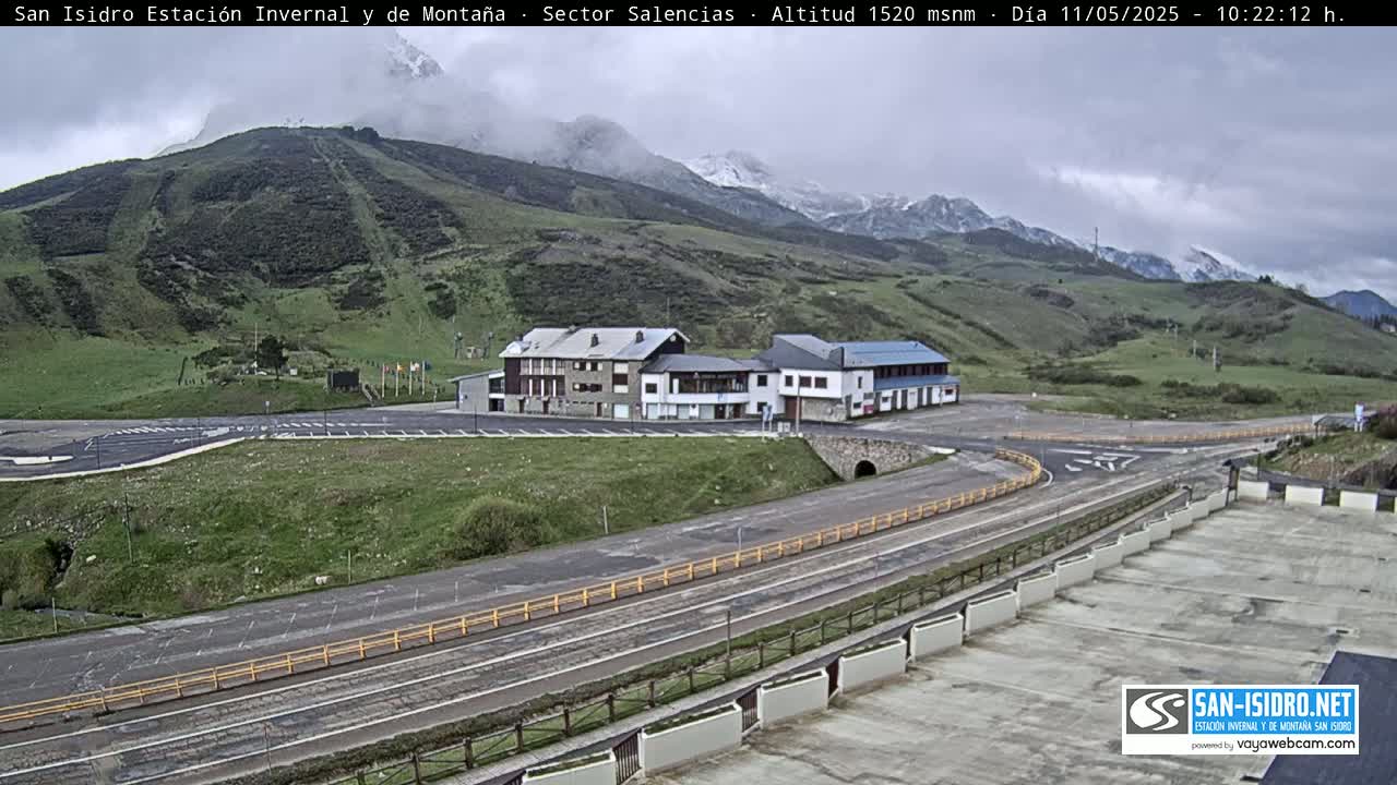 A mountain resort with several buildings sits beside a road curving through a grassy valley under an overcast sky.