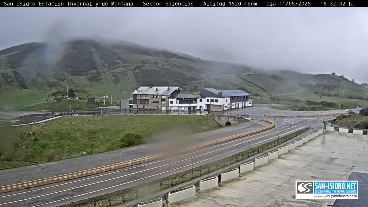 A misty mountain scene shows a roadway curving past a cluster of buildings in a parking lot.