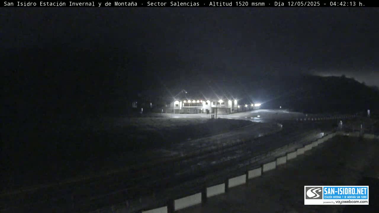 A brightly lit building sits in a dark, empty parking lot at night.