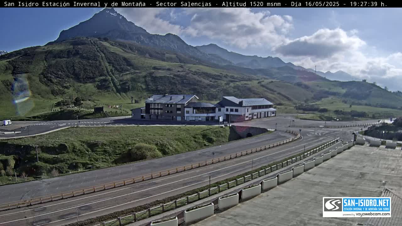 A mountainside building complex is visible from a high vantage point on a partly cloudy day, with roads and parking areas curving around it.