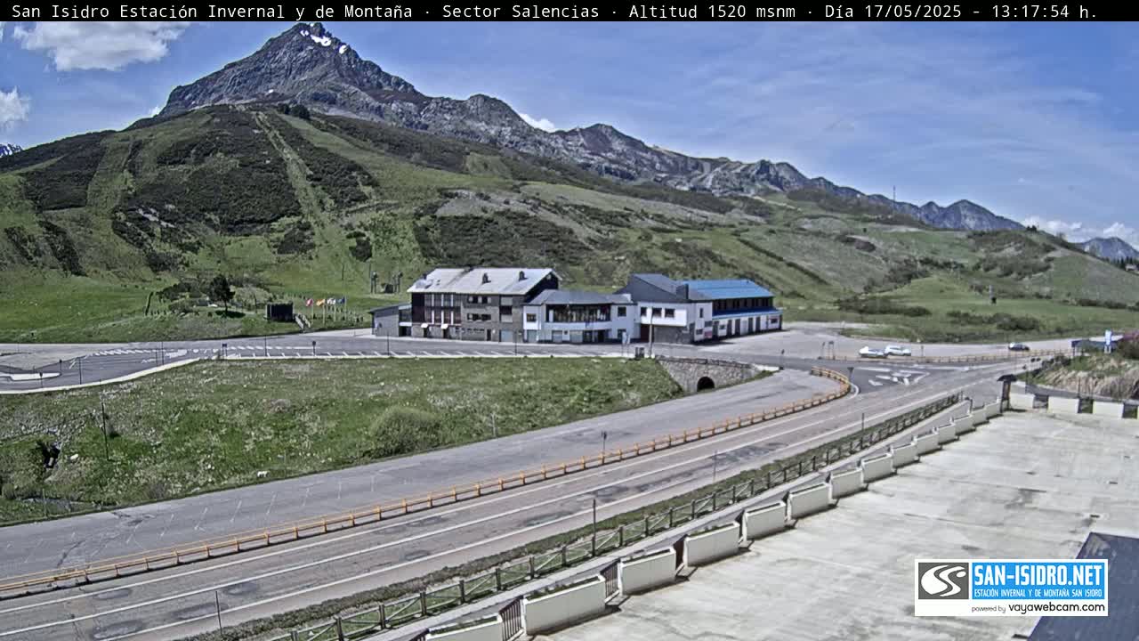 A mountainside parking lot and road are visible, with a few buildings, under a mostly sunny sky.