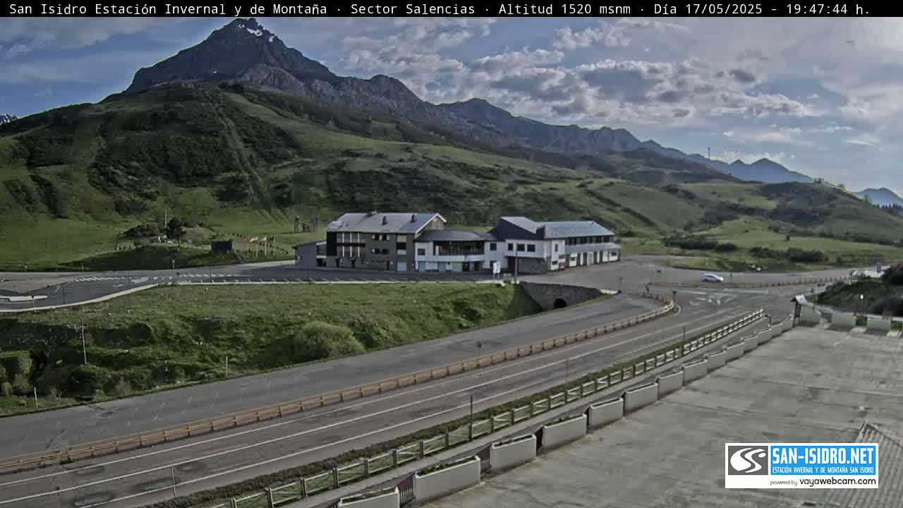 A mountain road winds past a building complex nestled in a valley, under a partly cloudy sky.