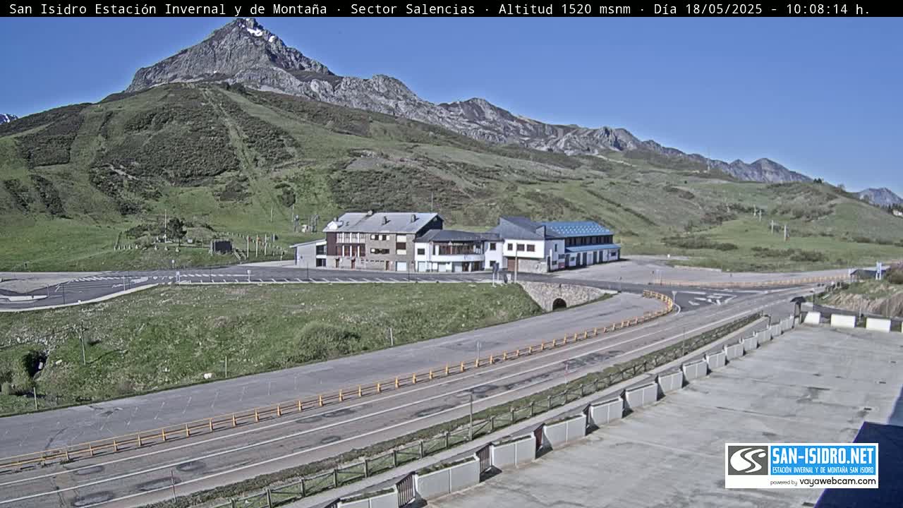 A mountainside resort with buildings and a parking lot is visible under a clear, sunny sky.