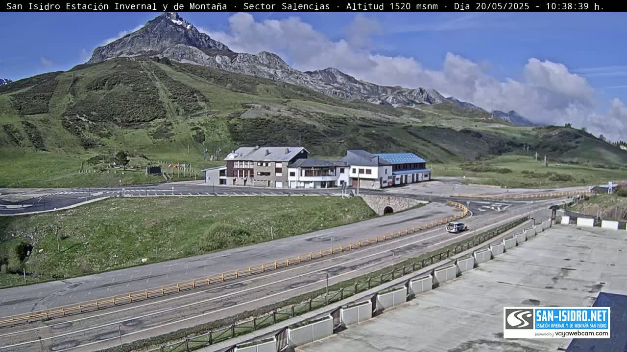 A mountainside parking lot with a few buildings and a road curves through it under a partly cloudy sky.