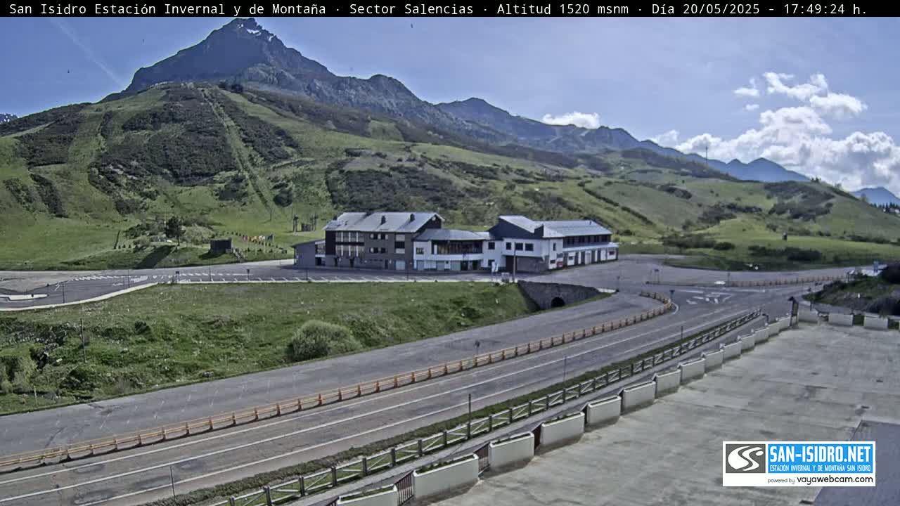 A mountainside building complex with parking lots and roads sits beneath a mostly clear sky, nestled among green hills and mountains.
