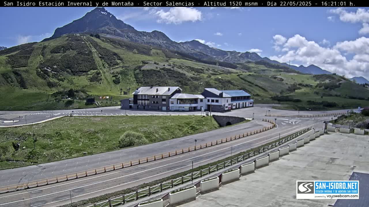 A mountainside building complex with a parking lot is visible, situated on a hillside road under a partly cloudy sky.