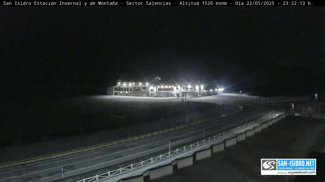 A brightly lit building sits atop a hill at night, overlooking a dark road and snow-covered ground.