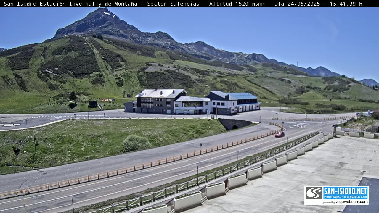 A mountain resort with a parking lot and buildings sits on a grassy hillside under a clear blue sky.