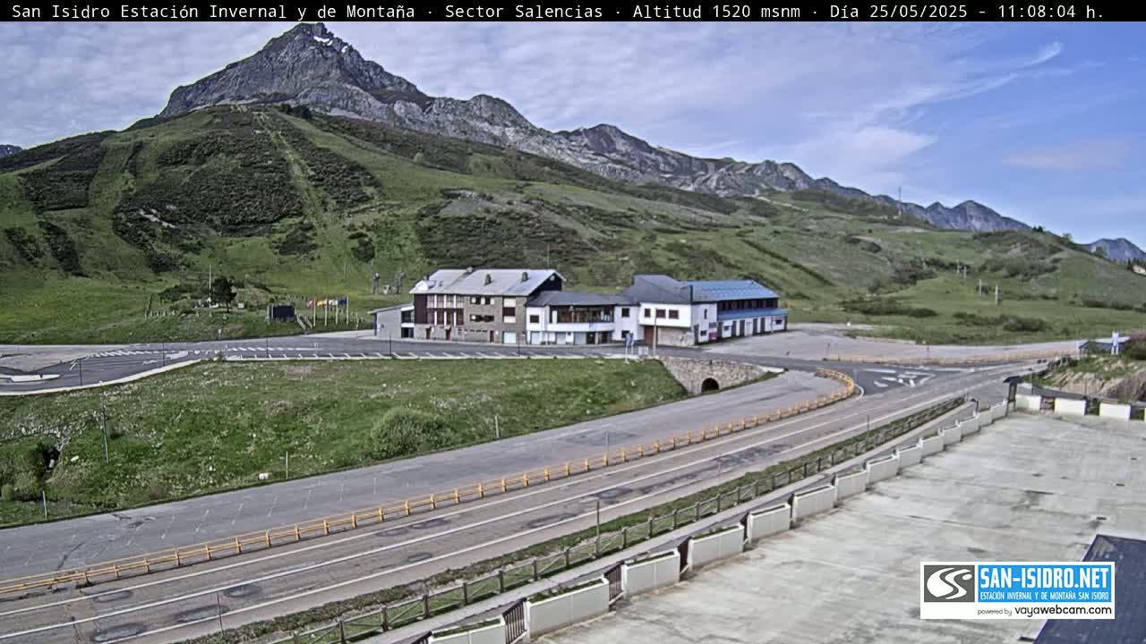 A mountainside parking lot with a few buildings, roads, and a mountain in the background under a mostly clear sky.
