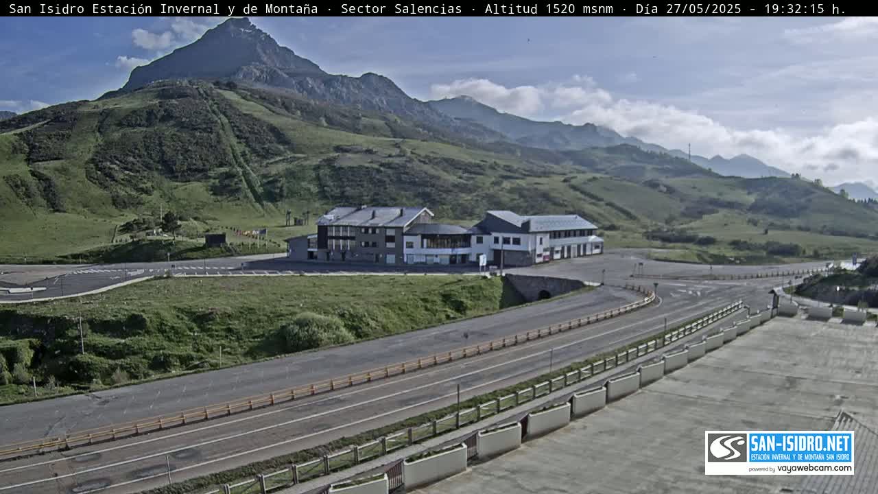 A mountainside building complex with a parking lot and roads is visible under a partly cloudy sky.