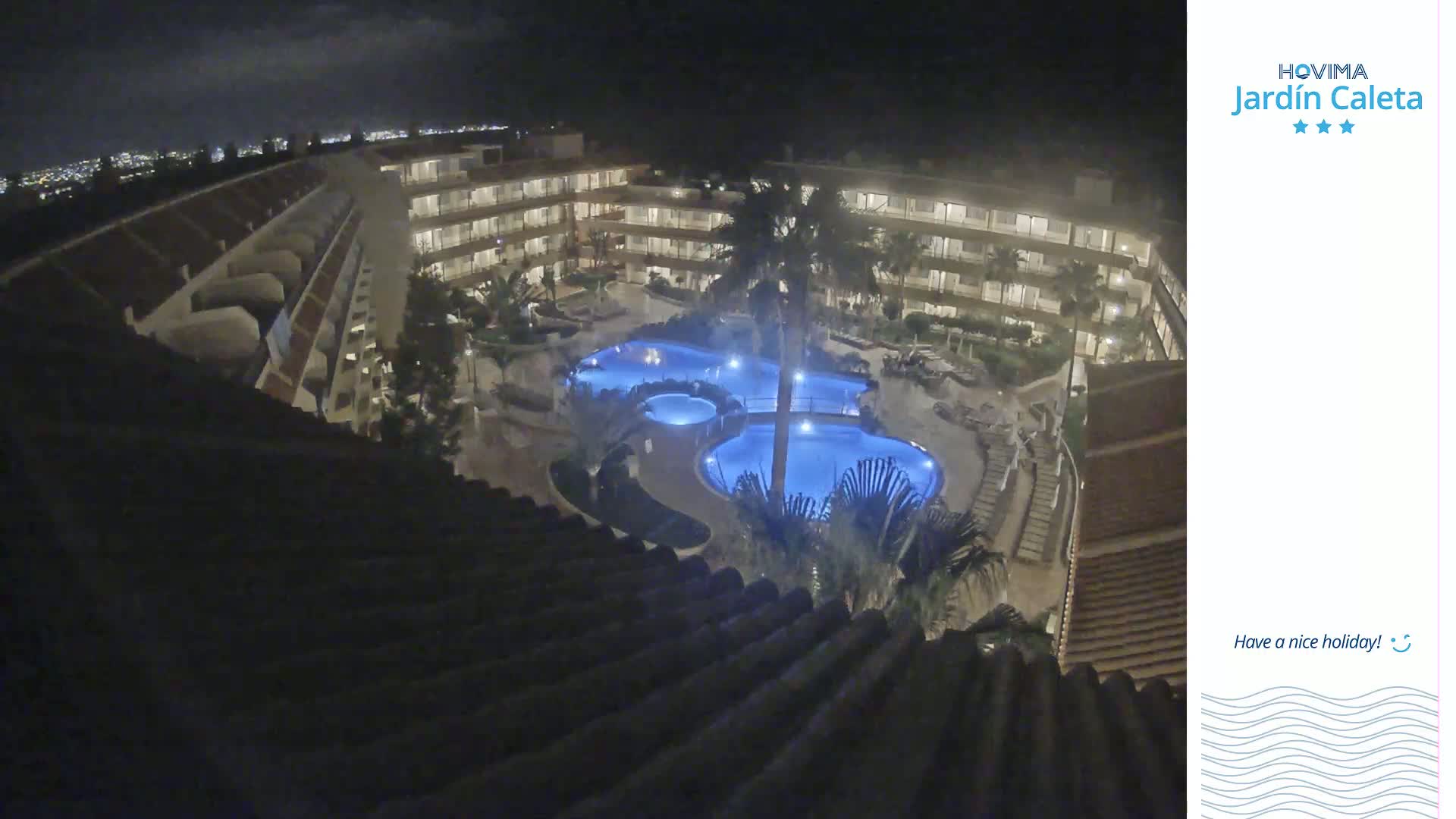 A nighttime view from a rooftop shows a multi-story building complex surrounding a lit swimming pool area.