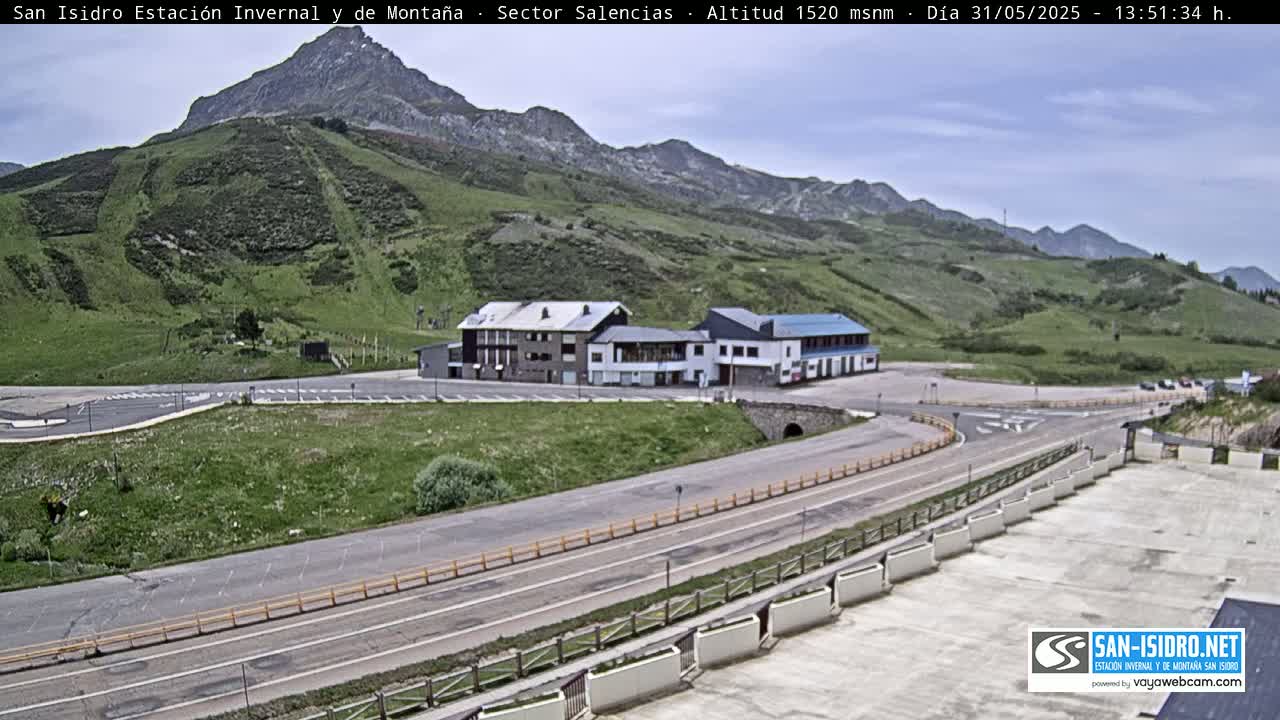 A mountainside parking lot and roadway with a few buildings are visible under a partly cloudy sky.