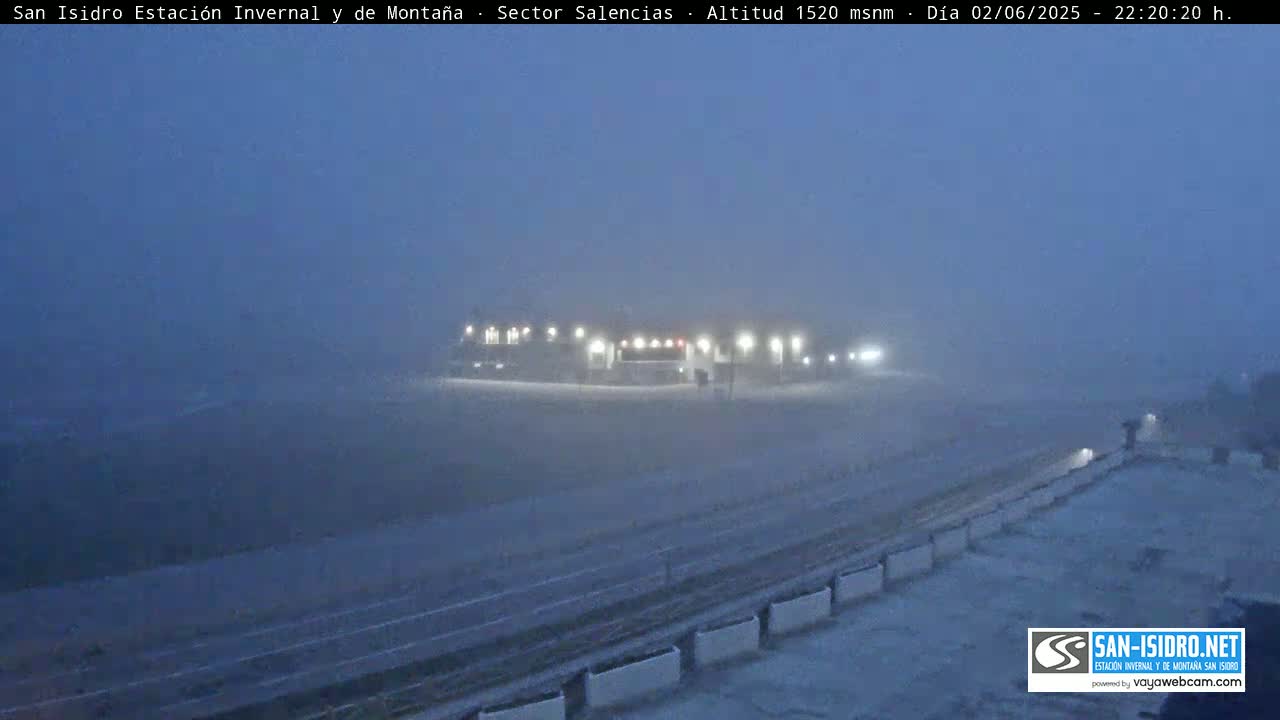 A snow-covered landscape is shrouded in heavy fog at night, with a dimly lit building visible in the distance.