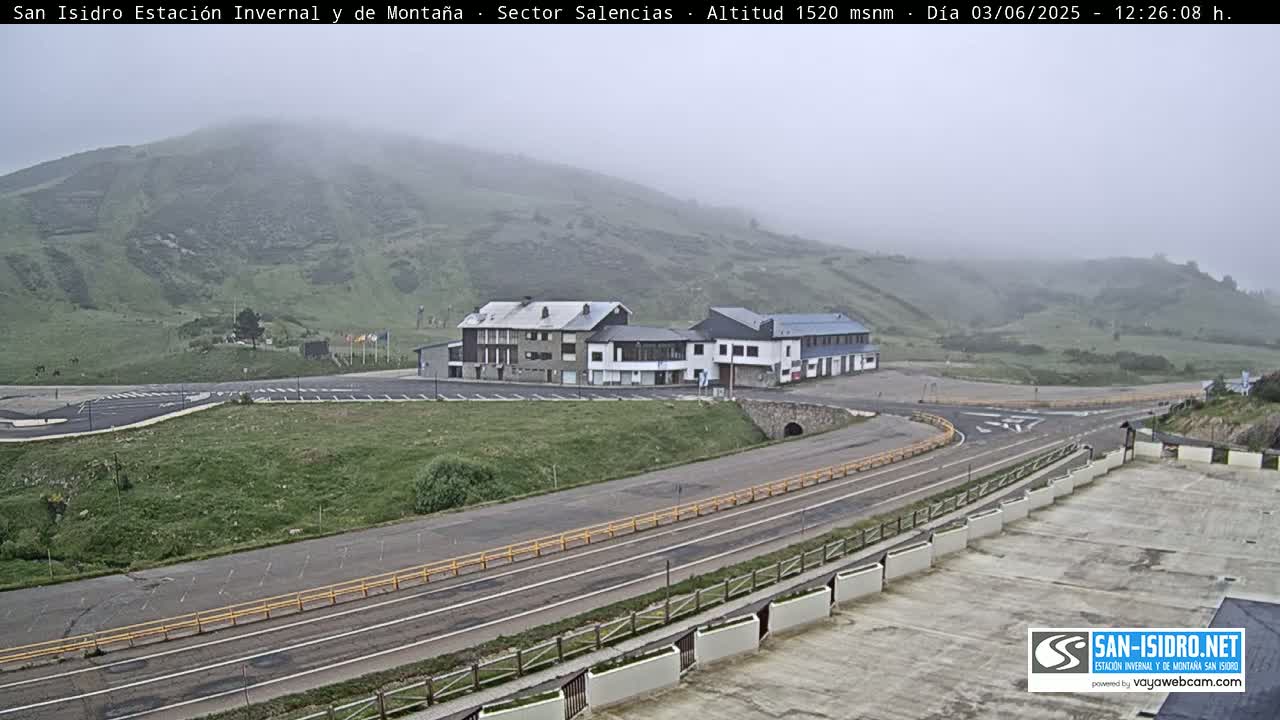A foggy mountain scene shows a roadway curving past a parking lot and several buildings, with additional parking areas visible in the foreground.