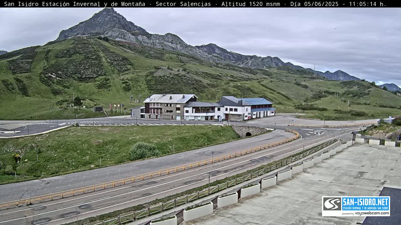 A mountain resort with several buildings sits beside a roadway that curves through a grassy landscape under a mostly cloudy sky.