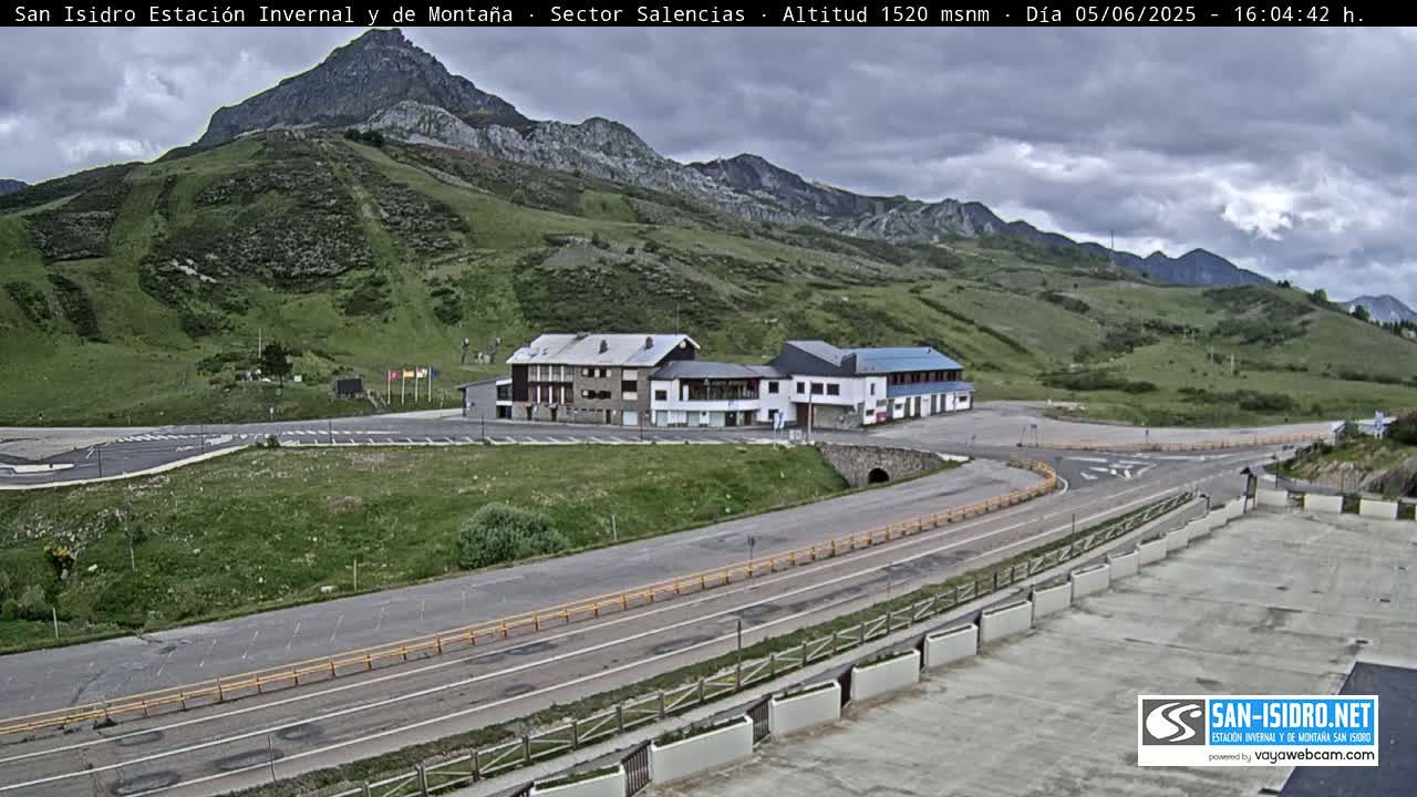 A mountainside parking lot with two buildings and a winding road under partly cloudy skies.
